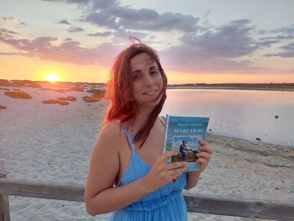 Io, il mio romanzo Mari Ermi e la spiaggia di Mari Ermi dietro di me nei colori del tramonto. 